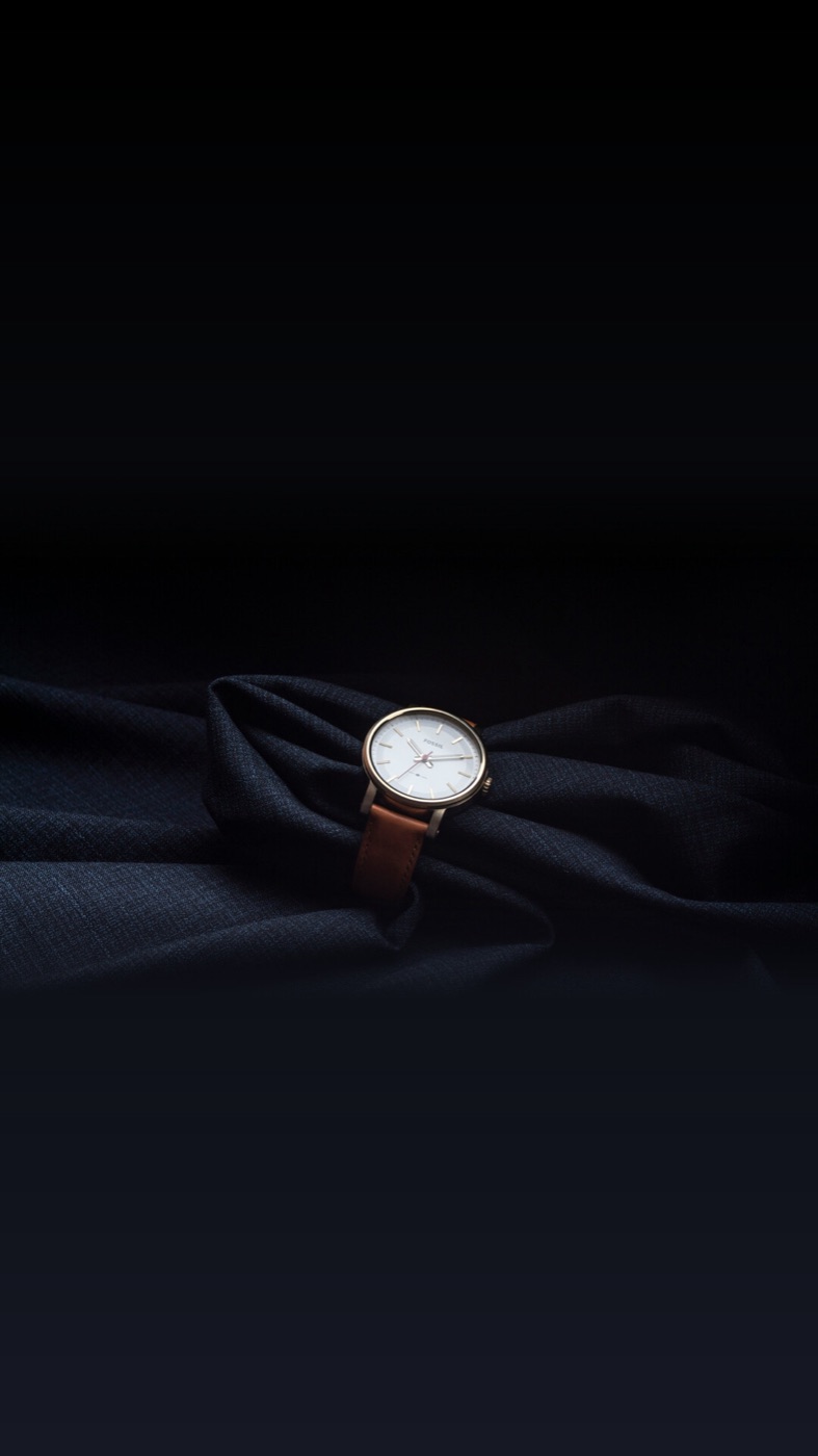 A white-dial watch on a dark leather strap, resting on folds of dark fabric in low light.